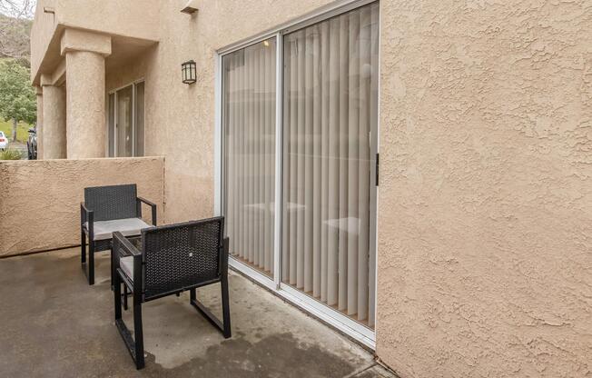 Outdoor patio area featuring two black chairs and a sliding glass door with vertical blinds. The wall is a textured beige, and the patio floor is concrete. Natural light is visible through the glass door, suggesting a bright environment.