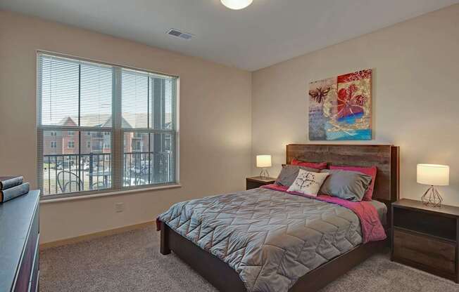 A bright bedroom here at Vue at Pinnacle Park with soft carpet, a large window with blinds and neighborhood views, a wood bed frame with gray and pink bedding, and matching nightstands with warm bedside lighting.