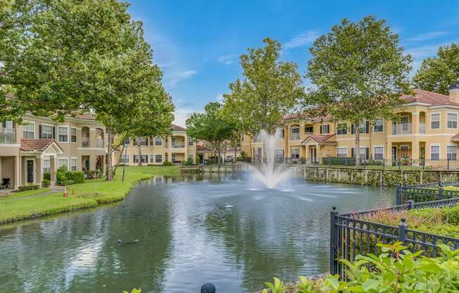 A fountain in the middle of a pond in front of apartment buildings.
