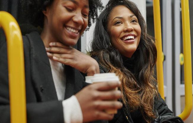 Two women are sitting on a bus and laughing while one of them holds a cup.