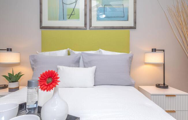 A cozy bedroom featuring a bed with gray and white pillows, a bright green headboard, and two bedside lamps. A small plant, a vase with a red flower, and a tray with a bottle and cups are arranged on a nightstand beside the bed. Abstract artwork adorns the wall above the headboard.