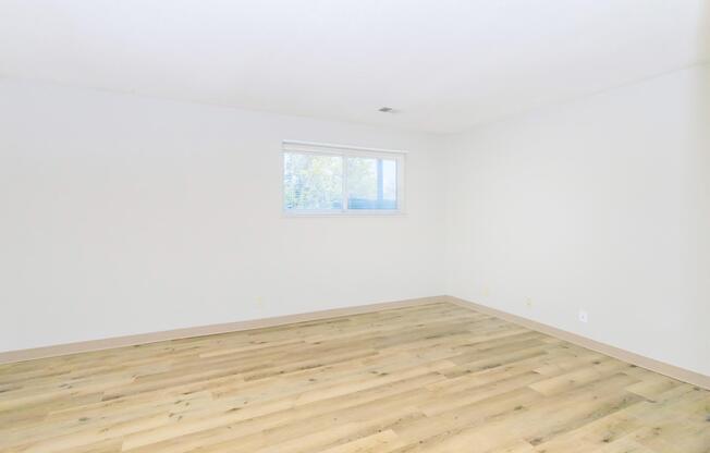 A spacious, empty room with light-colored walls and wooden flooring. A large window lets in natural light, illuminating the area. The overall atmosphere is bright and airy, suitable for various purposes such as a living space or office.