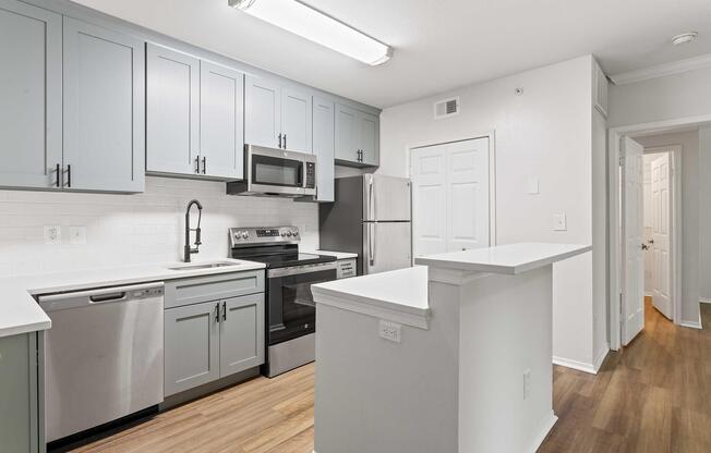 Modern kitchen featuring light gray cabinetry, stainless steel appliances, and a white tile backsplash. Includes a sink, dishwasher, microwave, and a large countertop that separates the kitchen from an adjoining hallway. The space has wood flooring and bright, overhead lighting.