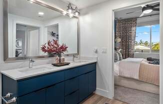 A bathroom with a large mirror, a sink, and a bedroom with a bed and a window.