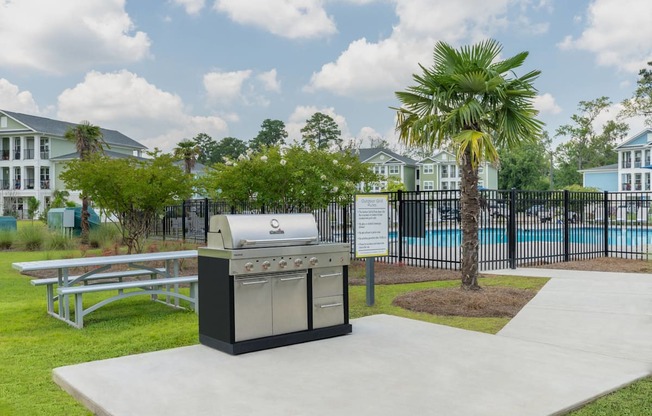 A BBQ grill is in the middle of a concrete area with a pool and palm tree in the background.