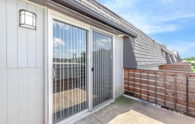 A view of a balcony with sliding glass doors leading outside. The doors have vertical blinds and a light fixture on the wall. The balcony has a wooden railing and a clear blue sky above, creating a bright and inviting atmosphere.
