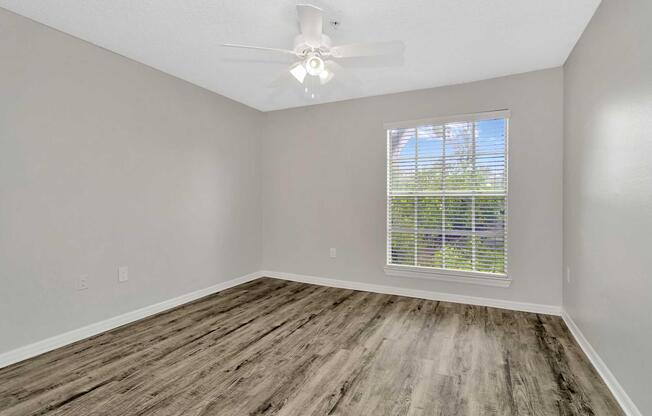 A clean, empty room featuring light gray walls, a ceiling fan, and a large window with white blinds that lets in natural light. The floor has a wood-like finish, and the space appears bright and inviting, ideal for customization or furnishing.
