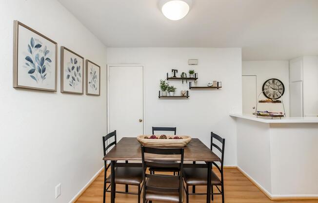 A modern dining area featuring a wooden table with a decorative bowl in the center. Four black chairs surround the table. On the wall, there are three framed botanical prints. A wall shelf displays plants and décor. In the background, a doorway leads to another room, and a clock is mounted on the wall.