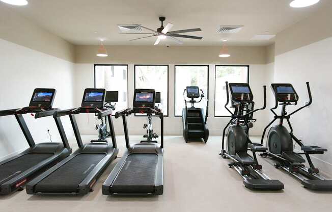 A row of treadmills and exercise bikes are lined up in a brightly lit gym.