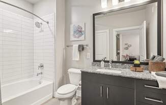 A bathroom with a white tub and shower, toilet, and granite countertop with a sink.