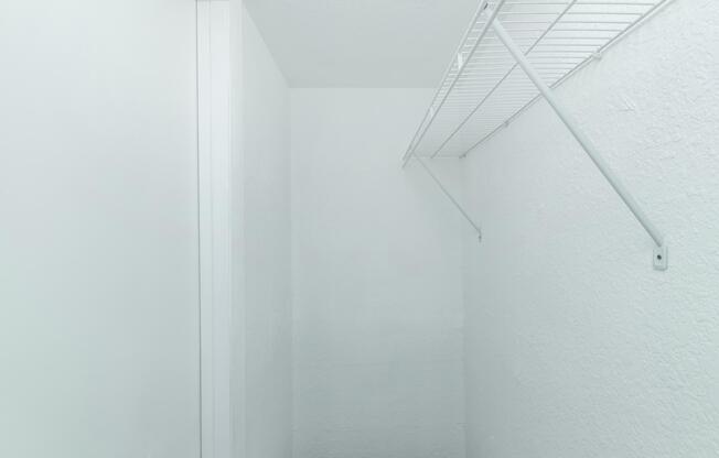 Empty white closet with bare walls and a wire shelving unit mounted high on one side. The space is well-lit and has a clean, minimalistic appearance, emphasizing its emptiness.