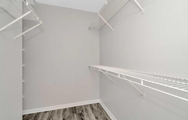 A spacious, empty closet featuring white wire shelves against a light gray wall. The floor is finished with a wood-like laminate, creating a clean, minimalist look. There are no items stored in the closet, emphasizing its openness and potential for organization.