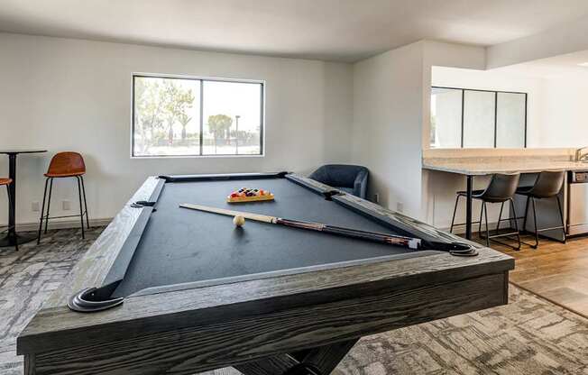 A pool table with a billiard cue and balls is in the foreground of a room with a dining table and chairs in the background.