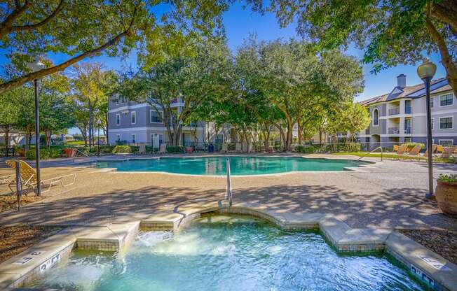 A pool and hot tub area at Saxony at Chase Oaks Apartments in Dallas, TX, with trees and apartment buildings in the background, offering a serene retreat for residents.