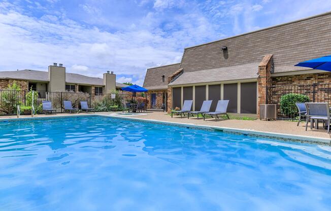 A clear blue swimming pool surrounded by lounge chairs and patio furniture, with umbrellas providing shade. The area is landscaped with greenery and brick structures in the background under a partly cloudy sky.
