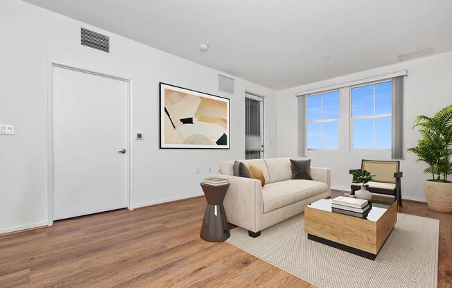 A modern, well-lit living room featuring a beige sofa with decorative pillows, a wooden coffee table with books and a small plant, a stylish side table, and a large piece of abstract wall art. There are two windows allowing natural light, and a potted plant in the corner. The flooring is wood, and the walls are painted white.