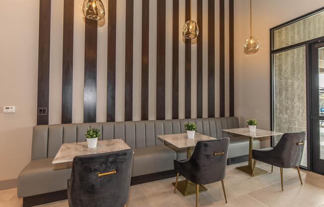 Interior of a modern cafe featuring a stylish design with black and white striped walls, two tables with marble tops, and green potted plants. Plush black chairs complement the decor, while pendant lights hang from the ceiling, creating an inviting atmosphere for customers.