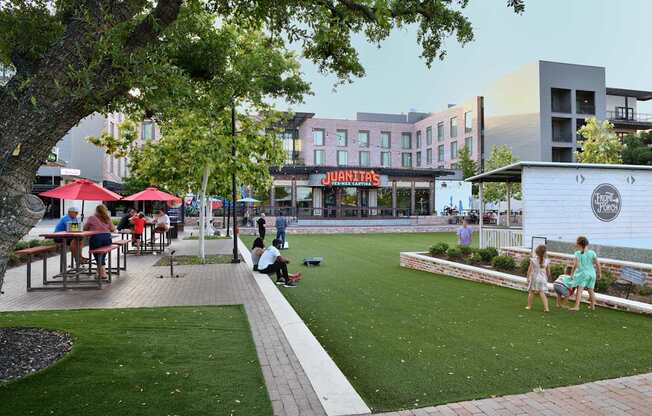 A group of people are sitting and standing on a grassy area in front of a building with a sign that says "JUANITAS".