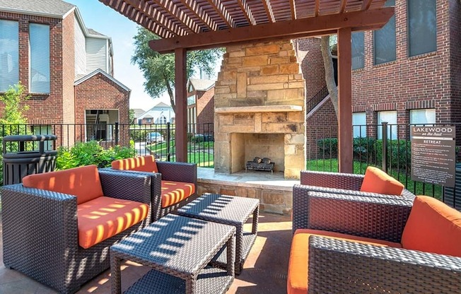 A cozy outdoor fireplace lounge here at Lakewood on the Trail with cushioned wicker seating, a stone fireplace, pergola shade, and fenced courtyard views, creating a warm and inviting gathering spot.
