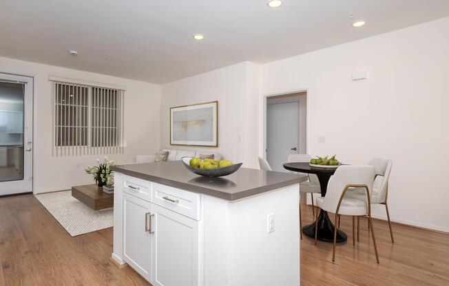 A modern, well-lit apartment interior featuring a kitchen island with a bowl of green apples, a dining area with a round table and white chairs, and a cozy living area with a sofa and artwork on the wall. Soft neutral colors and wooden flooring create a warm atmosphere.