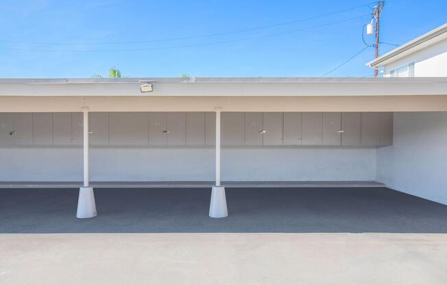 A clear view of a covered parking area with white poles supporting the roof. The ground is smooth and gray, and the walls are painted white. There are storage cabinets along the back wall and a bright blue sky visible above. The area appears clean and well-maintained, with no cars parked inside.