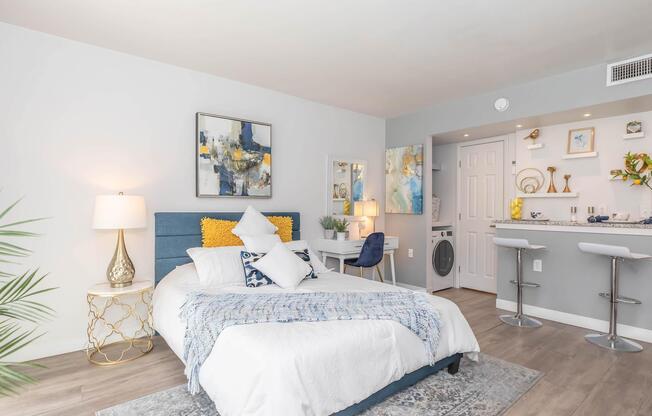 A cozy bedroom featuring a blue upholstered bed with white bedding and decorative pillows. There is a bedside table with a lamp, a small desk with a chair, and artwork on the walls. A laundry area is visible in the background, and the room has light wooden flooring and a modern, airy atmosphere.