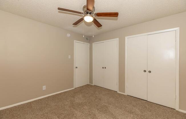 A small, empty room with light beige walls and a ceiling fan with wooden blades. The floor is covered in carpet, and there are two closed white closet doors on the right side. A single light fixture hangs from the ceiling, providing soft illumination. The room has a clean and simple appearance, suitable for various uses.