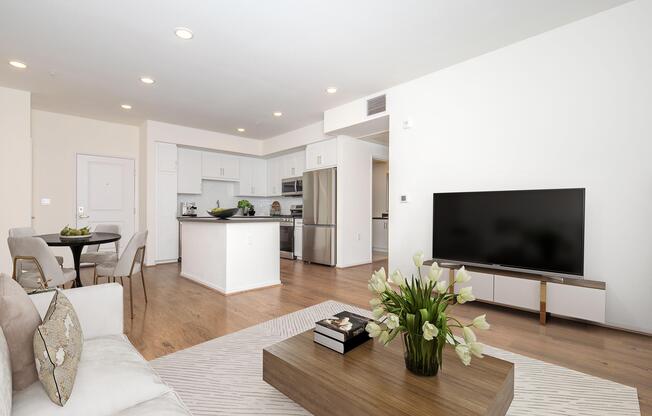 A modern living room and kitchen area featuring light-colored walls and wooden floors. The living room has a stylish sofa, a decorative coffee table with flowers, a large TV on a low cabinet, and a dining table with chairs in the background. Bright lighting enhances the open and inviting atmosphere.