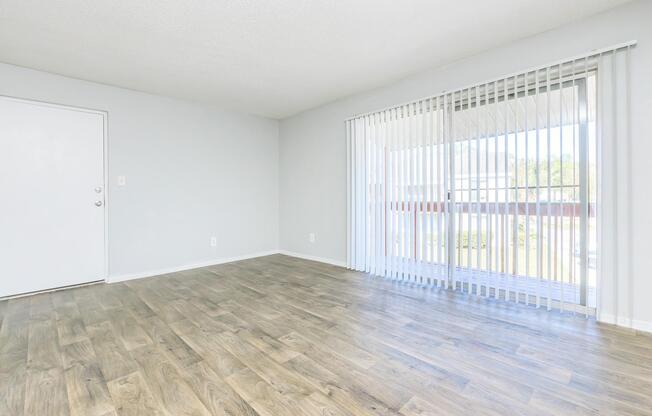 A spacious, empty room with light gray walls and wooden flooring. There is a closed white door on the left and large sliding glass doors with vertical blinds on the right, leading to a balcony. The space is well-lit with natural light.