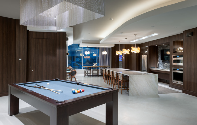 A clubhouse with dark wood-paneled walls, a billiards table under a rectangular chandelier with hanging silver chains, and a resident catering kitchen with two ovens, a large marble island with barstools, and an HDTV.