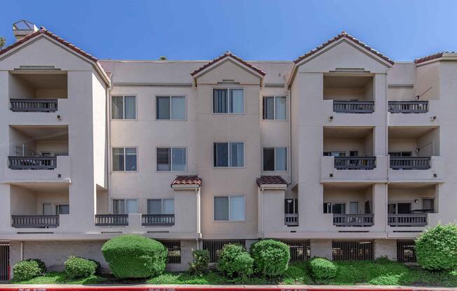 A three-story apartment building with a beige exterior, featuring balconies and large windows. The building is flanked by neatly trimmed green bushes. The sky is clear and blue, creating a bright background.