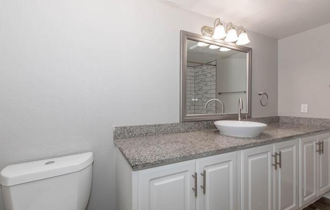 A modern bathroom featuring a white toilet, a gray granite countertop with a vessel sink, and a large mirror framed in silver. Wall-mounted lighting illuminates the space, which has neutral-colored walls and cabinetry, creating a clean and contemporary look.