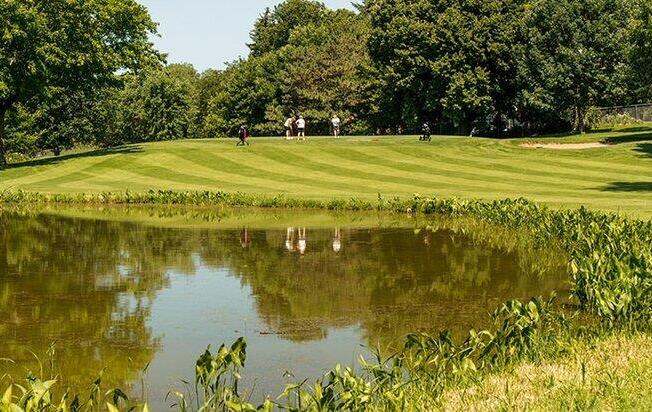 Sprawling green lawn and pond at a golf course on a bright, sunny day. Golfers can be seen in the distance among the trees.