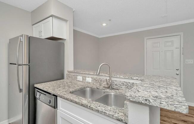 A modern kitchen with granite countertops, stainless steel appliances, including a refrigerator and dishwasher, and an open layout leading to a doorway. The walls are painted in a light neutral color, creating a bright and inviting atmosphere.