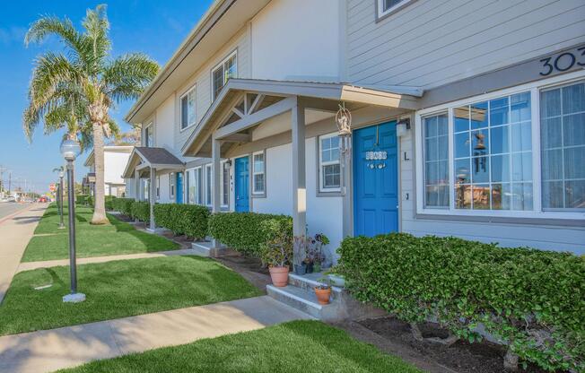 A row of well-maintained residential units with blue doors, surrounded by manicured shrubs and grass. Palm trees are visible nearby, and a sidewalk runs alongside the building, indicating a sunny day.