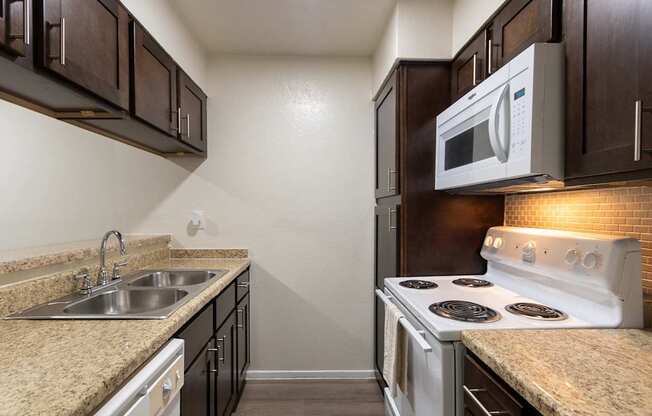 A kitchen with dark wood cabinets and white appliances in a 1 bedroom, 1 bath B, 629 sq ft apartment home at The Biltmore Apartments located in the Vickery Midtown neighborhood of Dallas, TX.