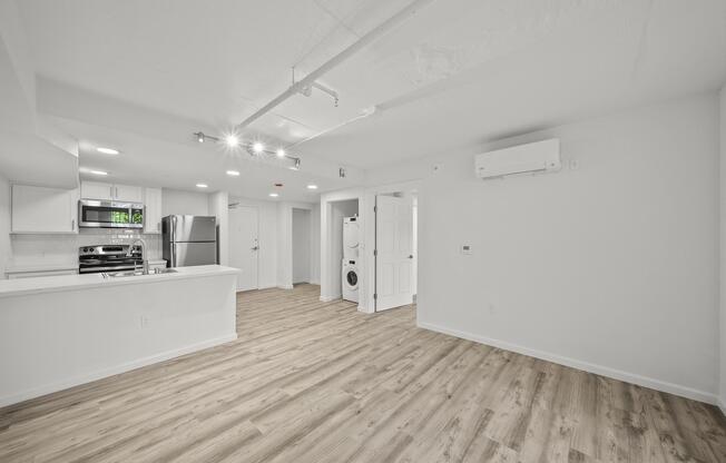 A spacious, modern interior of an apartment featuring light wood flooring, recessed lighting, and an open kitchen with stainless steel appliances. A washing machine is visible in a corner near a white door. The walls are painted in a light color, creating a bright and airy atmosphere.