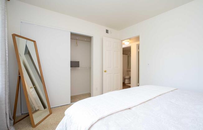 A well-lit bedroom featuring a neatly made bed with a white comforter. A large, leaning mirror is positioned against the wall, reflecting the room. Thereâs a closet door on the left, slightly open, and a bathroom door is visible in the background. The walls are painted white, creating a clean and spacious atmosphere.