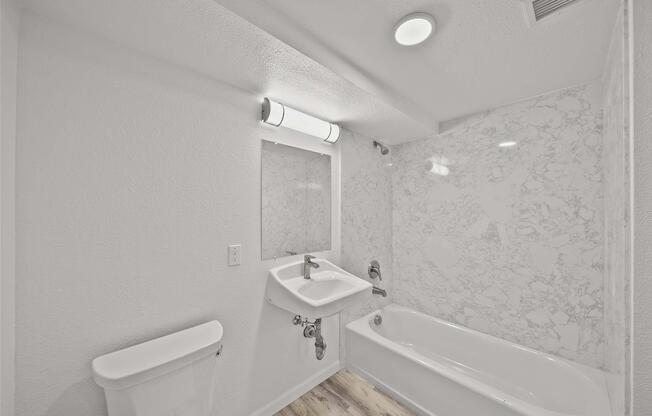 A modern bathroom featuring a white marble wall, a bathtub with a shower, a wall-mounted sink, a toilet, and overhead lighting. The space is well-lit and has neutral-colored walls and wood-style flooring.