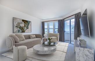 A modern living room featuring a round marble coffee table, a light gray sofa with decorative pillows, and two stylish chairs. There's a large piece of abstract art on the wall, and floor-to-ceiling windows offer a view outside. Soft natural light fills the room, creating a welcoming atmosphere.