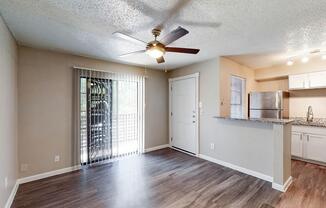 A cozy living room with a ceiling fan, featuring vinyl flooring and light-colored walls. A sliding glass door leads to a patio, allowing natural light. Adjacent to the living area is a kitchen with modern appliances, including a stainless steel refrigerator and granite countertops.