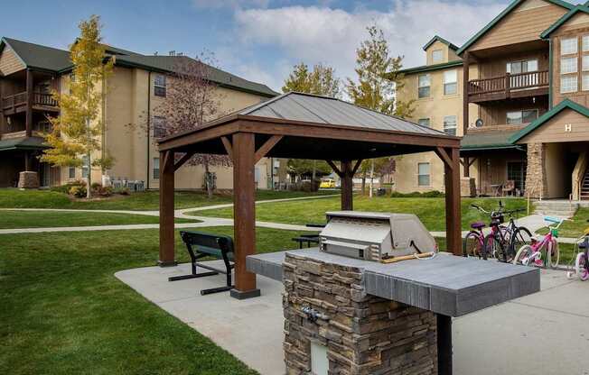 A covered picnic area with a grill in the middle of a grassy area in front of apartment buildings.