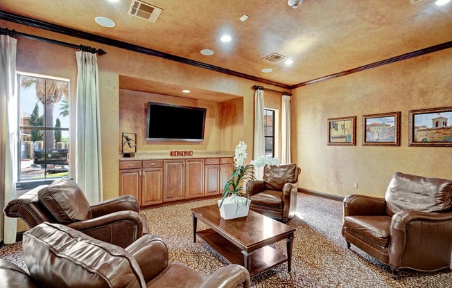 A cozy media lounge here at Tuscany at Faudree with plush leather seating arranged around a wooden coffee table. A large mounted television sits above built-in cabinetry, creating a dedicated entertainment area. Soft carpeting, framed wall art, and tall windows with curtains add warmth and natural light to the inviting community space.