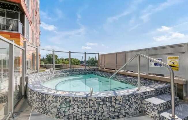 A tiled outdoor spa here at Blue and Lime featuring a circular design with mosaic tile walls, clear water, and built-in steps with metal handrails. Glass panels and privacy fencing surround the spa, while open sky and nearby buildings create a modern rooftop setting designed for relaxation and comfort.