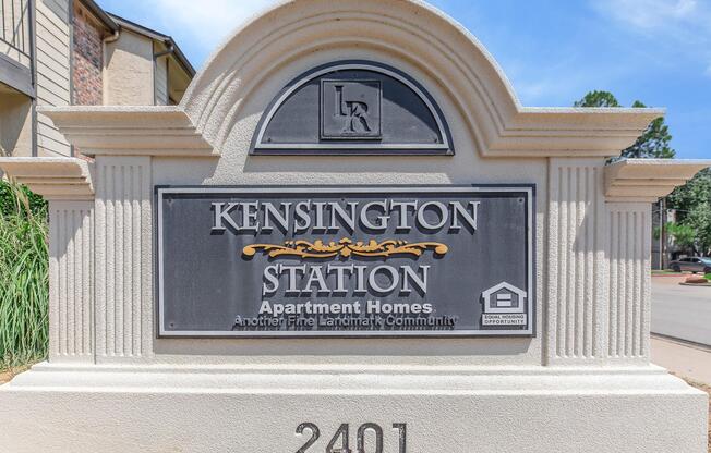 Sign for Kensington Station Apartment Homes, featuring elegant lettering and decorative elements. The sign indicates it's part of the Landmark Community, with the address 2401. The background has a clear blue sky and hints of green landscaping.