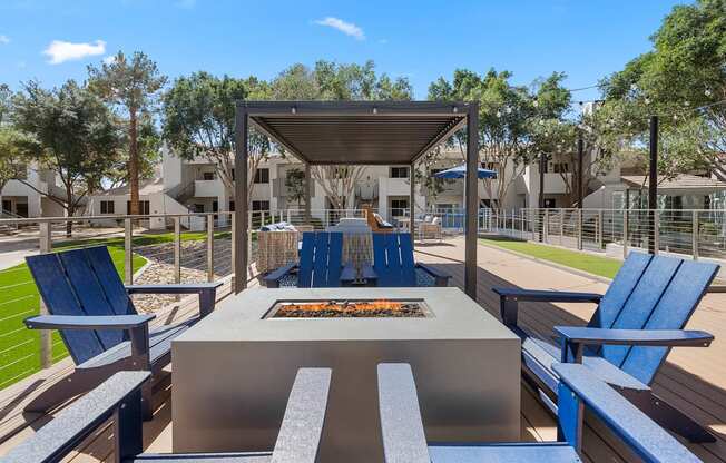 A fire pit is surrounded by blue chairs and benches in a sunny outdoor setting.