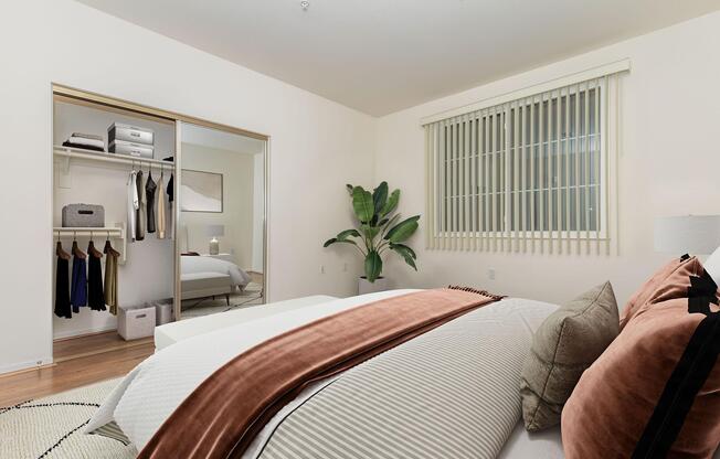 A stylish bedroom featuring a large bed with decorative pillows and a comforter in neutral tones. To the side, thereâs a closet with neatly hung clothes and storage boxes. A leafy plant adds a touch of greenery, and natural light filters through a window with blinds, creating a warm and inviting atmosphere.