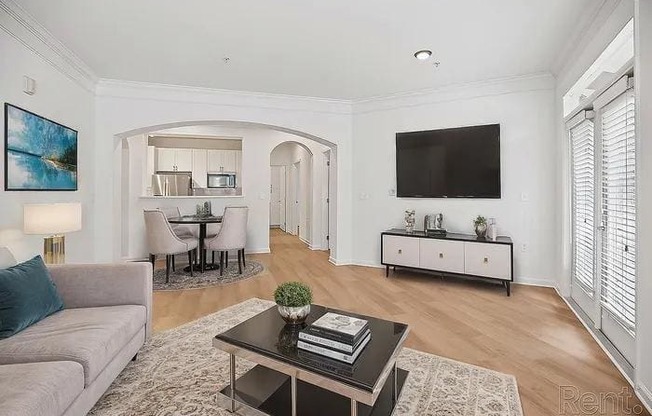 A living room with a grey couch, a black coffee table, a television, and a dining area in the background.