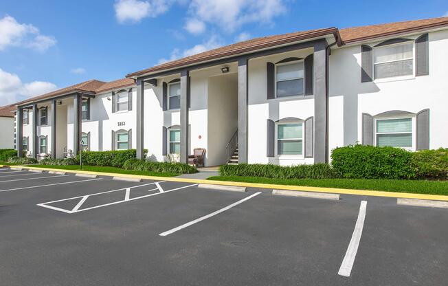 Two-story residential building with white exterior and dark window shutters. The entrance features a set of stairs leading to a covered porch. The surrounding area includes a well-maintained lawn and parked cars in a lot with clearly marked spaces. The sky is blue with some clouds.
