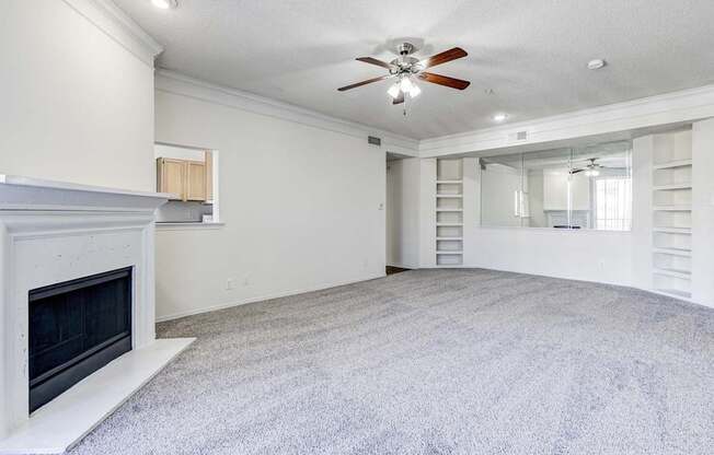 A large living room at Saxony at Chase Oaks Apartments in Dallas, TX, featuring built-in shelving, a ceiling fan, and carpeted floors, with a view of the dining and kitchen areas.
